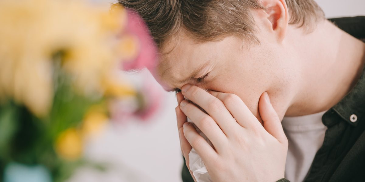 selective focus of man with pollen allergy covering face while sneezing in tissue near flowers